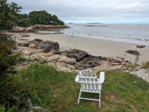 White Adirondack chair on a grassy bluff overlooking a quiet sandy beach and calm sea
