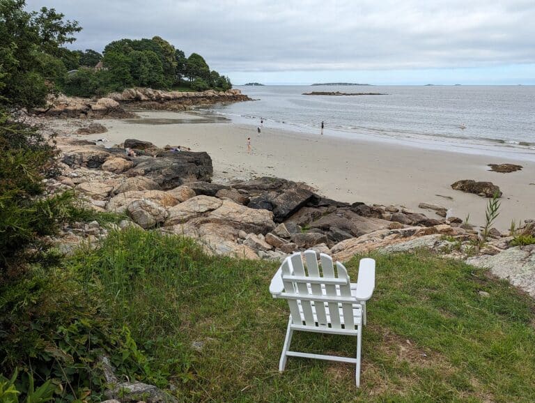 White Adirondack chair on a grassy bluff overlooking a quiet sandy beach and calm sea