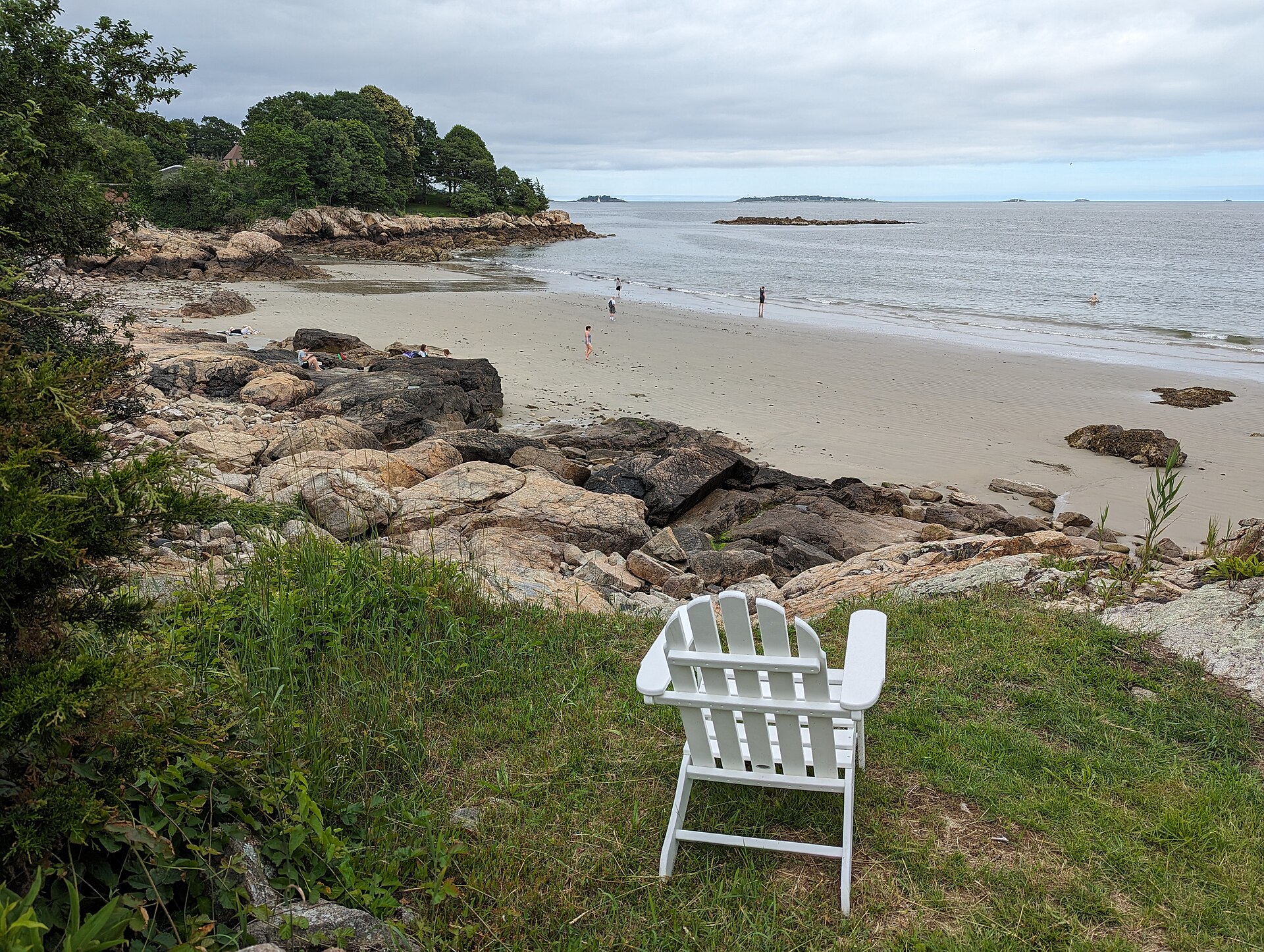 White Adirondack chair on a grassy bluff overlooking a quiet sandy beach and calm sea